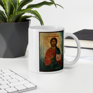 May include: White ceramic mug featuring a religious icon. The artwork depicts a figure with a halo, holding a book with Greek letters. The figure is wearing red and green robes. The mug is on a white desk with a keyboard and a plant.