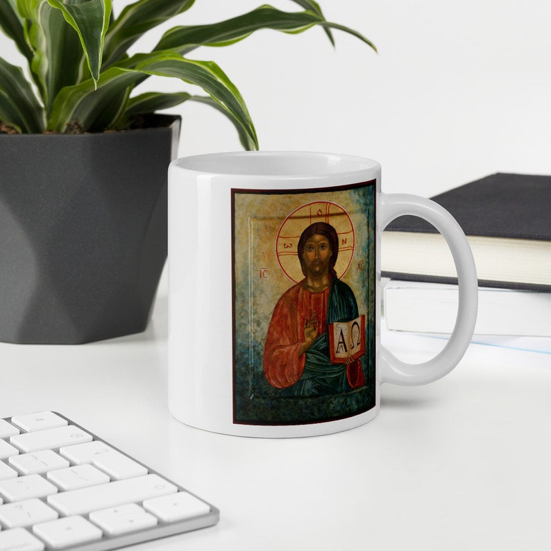 May include: White ceramic mug featuring a religious icon. The artwork depicts a figure with a halo, holding a book with Greek letters. The figure is wearing red and green robes. The mug is on a white desk with a keyboard and a plant.