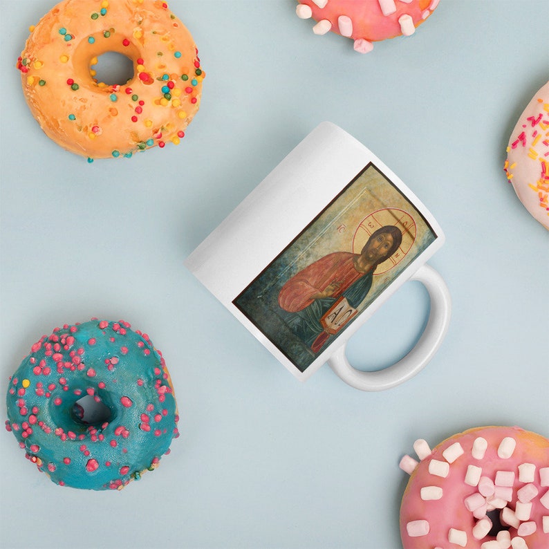 May include: White ceramic mug featuring a religious icon illustration. The mug is surrounded by colorful donuts with sprinkles on a light blue surface. The icon depicts a figure with a halo, holding a book.