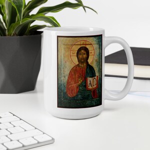 May include: White ceramic mug featuring a religious icon of a figure in red and green robes, holding a book with Greek letters. The icon is set against a gold background with a halo. The mug is on a white surface.