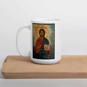 May include: White ceramic mug featuring a religious icon. The icon depicts a figure in red and blue robes holding a book with Greek letters. The mug is on a wooden surface.