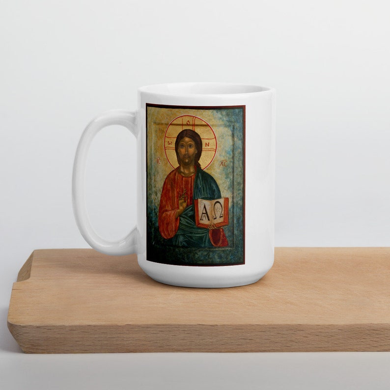 May include: White ceramic mug featuring a religious icon. The icon depicts a figure in red and blue robes holding a book with Greek letters. The mug is on a wooden surface.