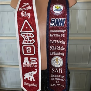 May include: A red and white graduation sash with the text "Zion Danielle Riley", "A&Xi; SPRING '20", "47", and "Divergent Soul". A blue and white graduation sash with the text "BNN", "Henderson Davis Player. Miss '18-'19 Pres. '19 '21", "TMCF Scholar", "SCBA Scholar", "A. Williams Scholar", "&Sigma;&Alpha;&Pi;", "Comm. Club", "S.O.L. Parliamentarian 20-21", and "M.A.C VP '17-'18".