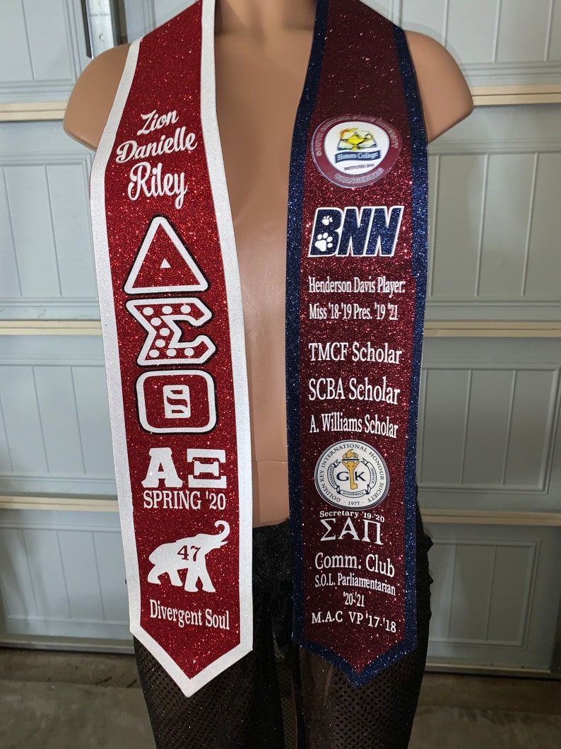 May include: A red and white graduation sash with the text "Zion Danielle Riley", "A&Xi; SPRING '20", "47", and "Divergent Soul". A blue and white graduation sash with the text "BNN", "Henderson Davis Player. Miss '18-'19 Pres. '19 '21", "TMCF Scholar", "SCBA Scholar", "A. Williams Scholar", "&Sigma;&Alpha;&Pi;", "Comm. Club", "S.O.L. Parliamentarian 20-21", and "M.A.C VP '17-'18".