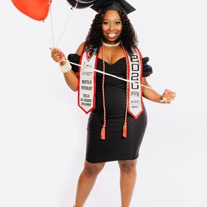 May include: A woman in a black dress and graduation cap holds a white and red graduation sash with the text "Crenshaw University Master of Psychology Child and Adolescent Development 2020" and red tassels. She is holding balloons in her left hand.