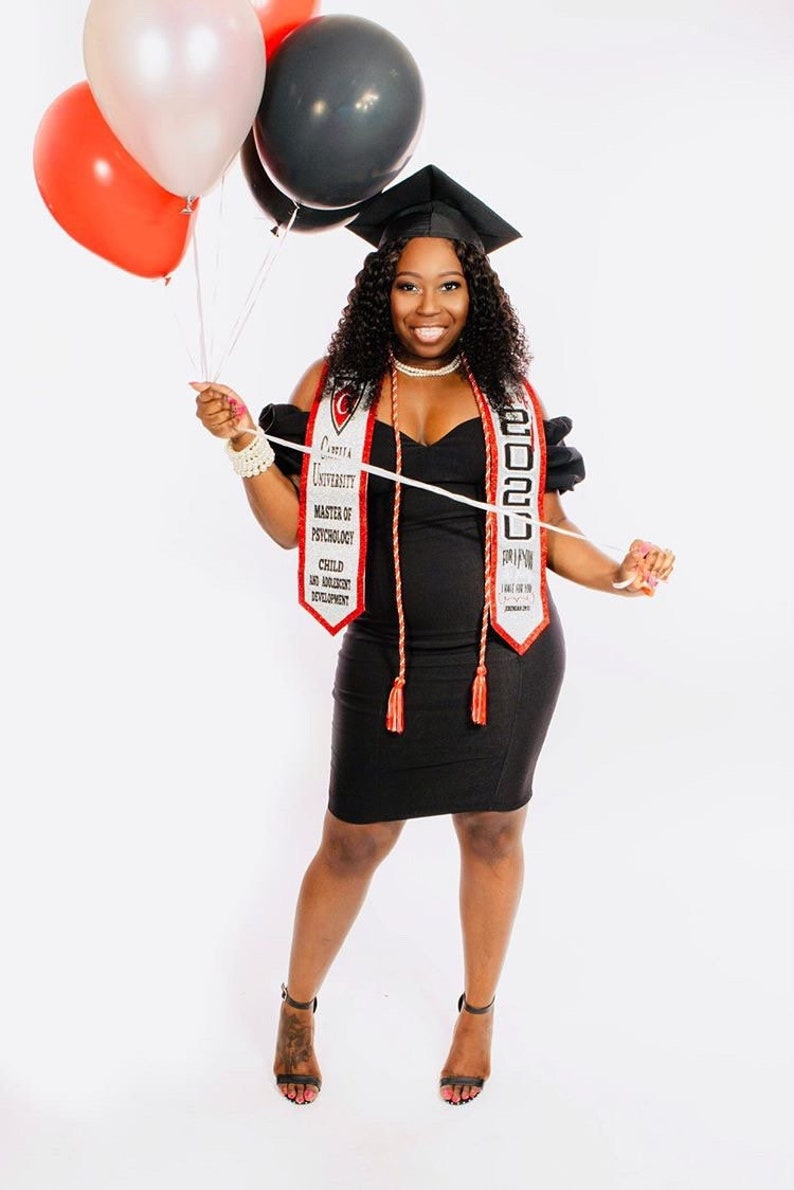May include: A woman in a black dress and graduation cap holds a white and red graduation sash with the text "Crenshaw University Master of Psychology Child and Adolescent Development 2020" and red tassels. She is holding balloons in her left hand.