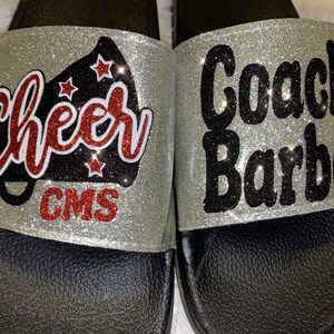 May include: A pair of silver glitter slide sandals with black straps. One sandal has the text "Cheer" with a red megaphone and the text "GMS" in red. The other sandal has the text "Coach Barber" in black.