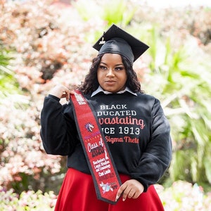 May include: A woman wearing a black graduation cap and gown, a red skirt, and a black sweatshirt with the text "Black Educated and Vastating Since 1913" and "Class of 2020" and "Sigma Theta". She is holding a red and black graduation sash with the text "Paul Quinn College" and "Bachelors in Legal Studies and Criminology Cum Laude".