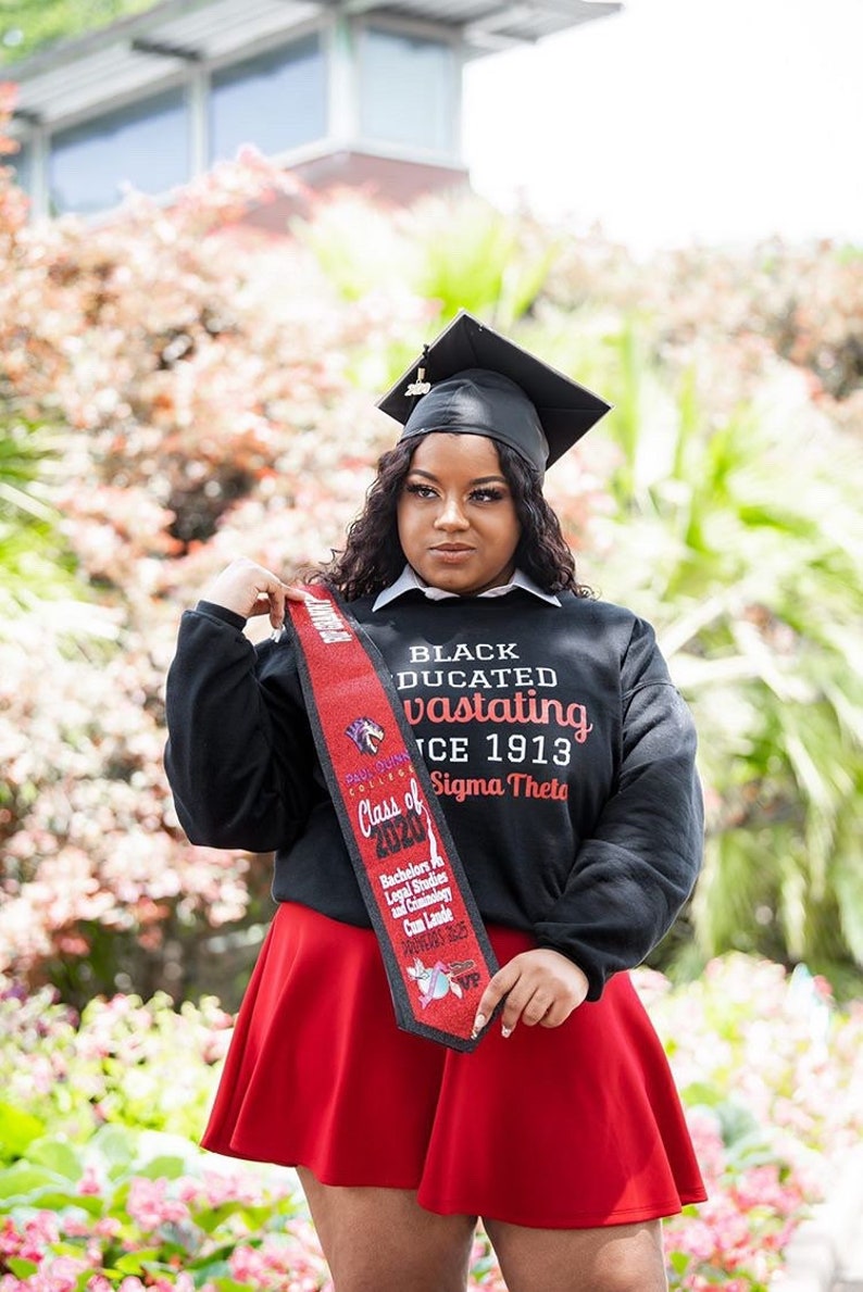 May include: A woman wearing a black graduation cap and gown, a red skirt, and a black sweatshirt with the text "Black Educated and Vastating Since 1913" and "Class of 2020" and "Sigma Theta". She is holding a red and black graduation sash with the text "Paul Quinn College" and "Bachelors in Legal Studies and Criminology Cum Laude".