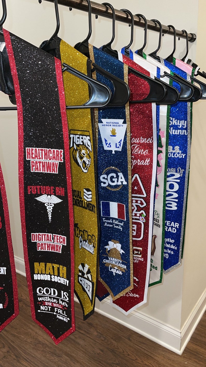 May include: A collection of graduation sashes with various colors and designs, including gold, red, blue, black, and white. The sashes feature text such as "Healthcare Pathway", "Future RN", "Digital Tech Pathway", "Math Honor Society", "God is within her, she will not fall", "Tigers", "Dual Enrollment", "SGA", "Varsity Cheerleader Captain", "Tournei Tena Pratt", "Skyy Vunn", "BS. Biology", "Class of 2023", and "Year Rad".