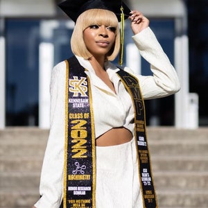 May include: A woman wearing a white graduation gown and a black cap with gold trim. She is holding a black and gold graduation sash with the text "Kennesaw State" and "Class of 2022".