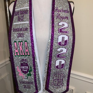May include: Silver graduation stole with purple trim and white lettering. The stole features the text "Aysheeda Rogers", "2020", "AKA", "Greek Council", "Resident Assistant 2018-2020", and "Orientation Leader 2018".