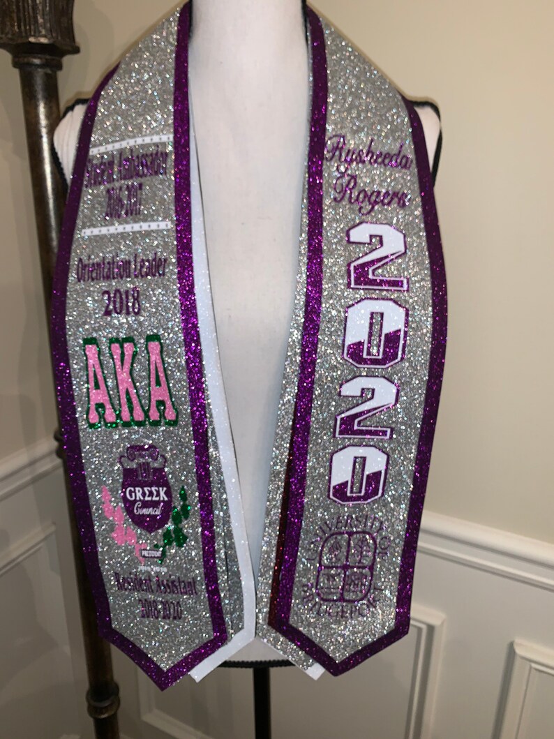May include: Silver graduation stole with purple trim and white lettering. The stole features the text "Aysheeda Rogers", "2020", "AKA", "Greek Council", "Resident Assistant 2018-2020", and "Orientation Leader 2018".