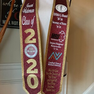May include: A maroon and gold graduation stole with the text "Imani Wideman Class of 2020" and the Florida State University seal. The stole also features several patches, including "SNMA L-Board '18-'20 Secretary of Points 19-20", "MRU Emergency Management Responder 17-20", and "Certified Clinical Medical Assistant". The stole has a quote from Psalms 20:4, "May the Lord Grant You the Desire of Your Heart and Make All Your Plans Succeed."