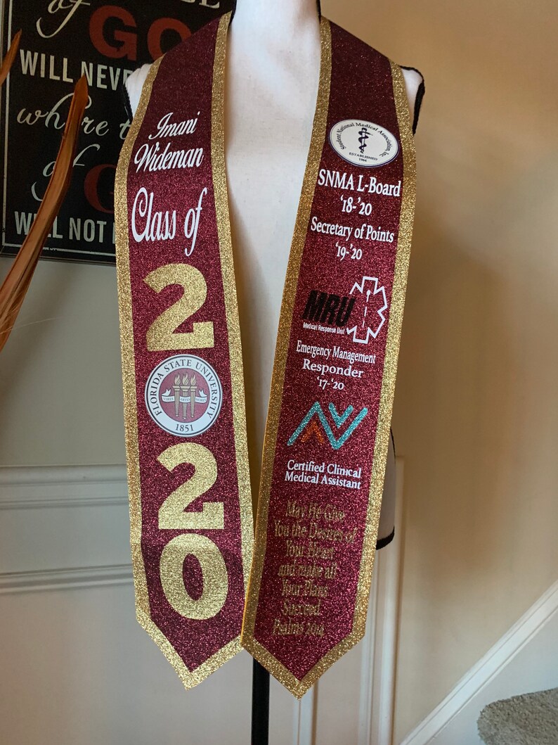 May include: A maroon and gold graduation stole with the text "Imani Wideman Class of 2020" and the Florida State University seal. The stole also features several patches, including "SNMA L-Board '18-'20 Secretary of Points 19-20", "MRU Emergency Management Responder 17-20", and "Certified Clinical Medical Assistant". The stole has a quote from Psalms 20:4, "May the Lord Grant You the Desire of Your Heart and Make All Your Plans Succeed."