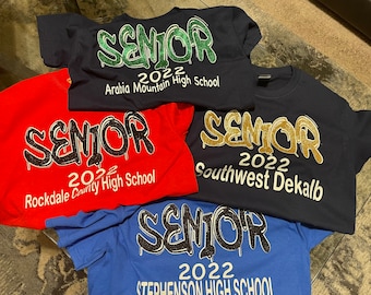 May include: Four different t-shirts with the word "Senior" in a graffiti style font. Each shirt has a different school name and the year 2022. The shirts are red, blue, navy blue, and royal blue.