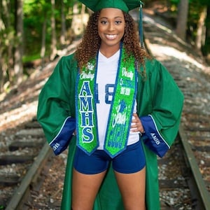 May include: A person wearing a green graduation gown with a blue and green sash that says "A.M.H.S. Senior Sterling" and "Varsity Volleyball 2017-2020". The person is also wearing a white jersey with the number "18" on it, blue shorts, and black knee-high socks.