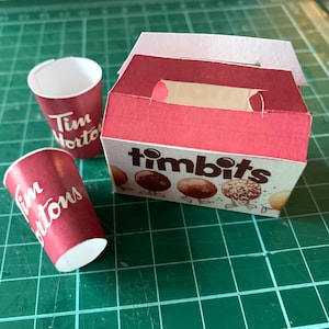 Puede incluir: Una caja roja y blanca de Tim Hortons con el texto "timbits" en la parte delantera. Hay dos vasos rojos de Tim Hortons junto a la caja.