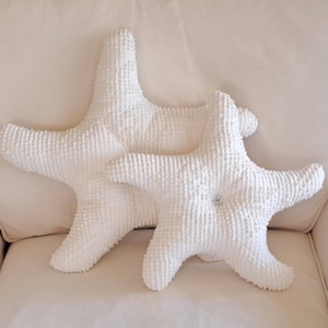 Puede incluir: Dos cojines decorativos blancos con forma de estrella de mar y superficies texturizadas. Los cojines están colocados sobre un sofá color crema. Un cojín tiene un botón en el centro. Decoración costera.