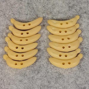 Könnte beinhalten: Eine Ansammlung von zwölf bananenförmigen Knöpfen, die in zwei vertikalen Spalten angeordnet sind. Jeder Knopf ist in einem einheitlichen Hellgelb gehalten, hat eine glatte, geschwungene Oberfläche und zwei kleine Löcher in der Mitte.