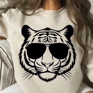 Könnte beinhalten: Ein cremefarbener Pullover mit einem schwarzen Aufdruck eines Tigers mit Sonnenbrille. Die Augen des Tigers sind geschlossen und die Sonnenbrille ist rund.