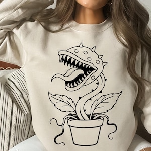 以下が含まれることがあります： ベージュのクルーネックトレーナーで、鉢植えの食虫植物の黒い線画が描かれています。植物は鋭い歯のある大きく開いた口、長い舌、頭部のトゲが特徴で、衣服の前面中央に配置されています。