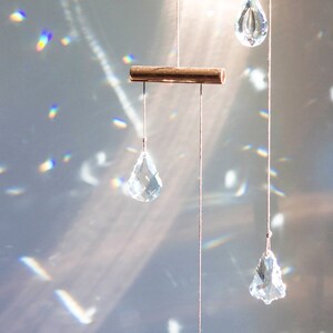May include: A crystal suncatcher with multiple clear crystal drops hanging from a copper bar. The suncatcher is hanging from a ceiling and casting rainbows on the wall.
