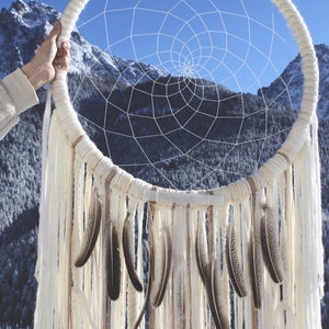 Puede incluir: Un atrapasueños blanco con plumas marrones que cuelgan de la parte inferior. El atrapasueños está frente a una cordillera nevada con un cielo azul.