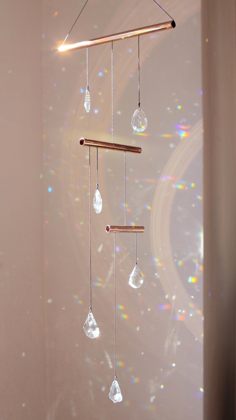 May include: A copper crystal suncatcher with five clear crystal drops hanging from three copper bars. The suncatcher casts rainbows on the wall behind it.