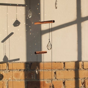 May include: A copper and crystal mobile hanging from a ceiling with a brick wall in the background. The mobile has three copper bars and four crystal teardrop shapes.