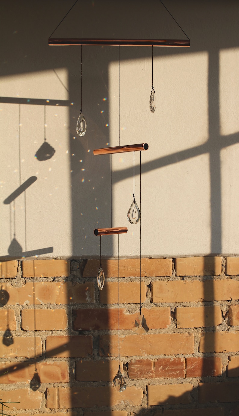 May include: A copper and crystal mobile hanging from a ceiling with a brick wall in the background. The mobile has three copper bars and four crystal teardrop shapes.