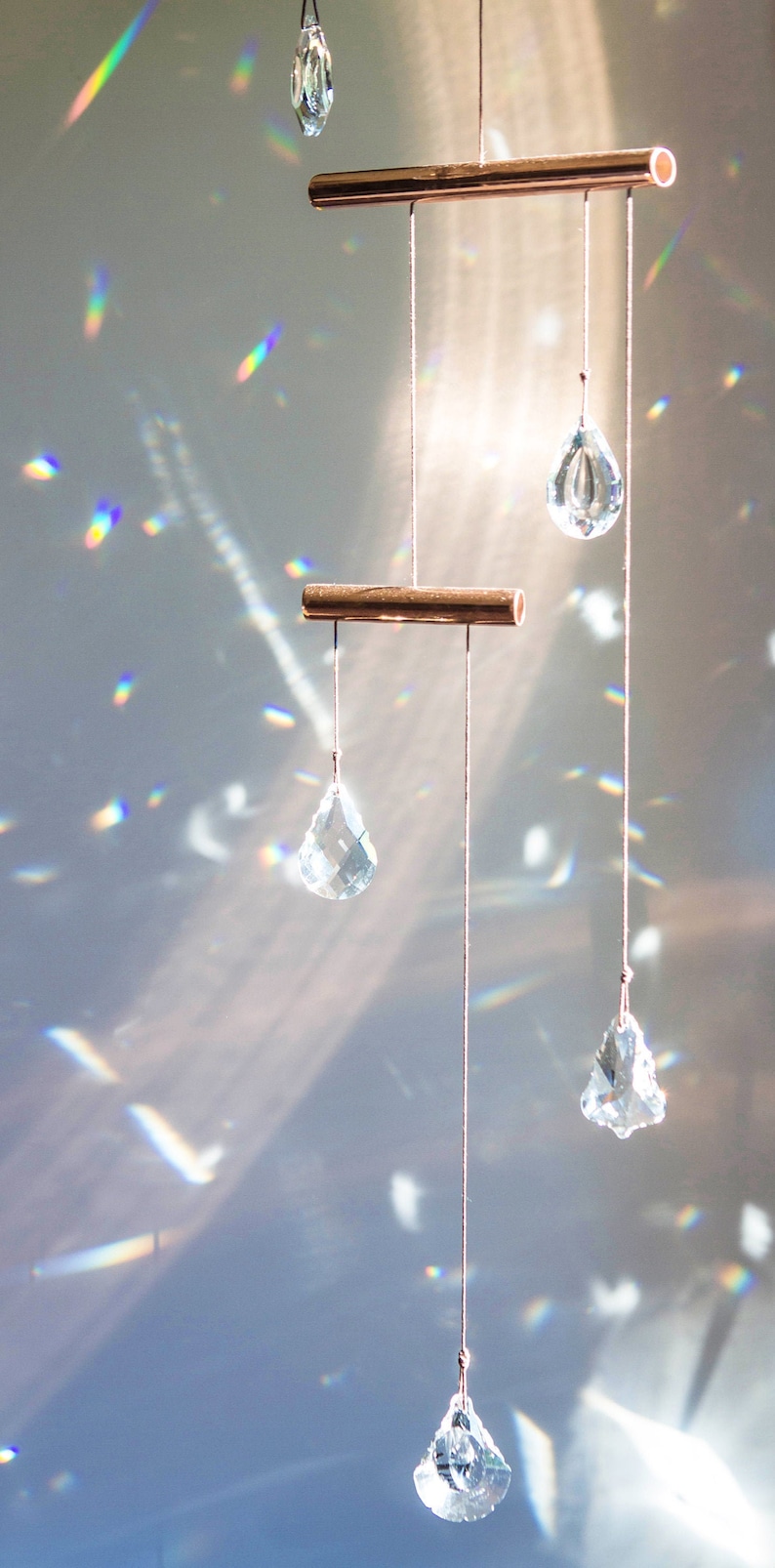 May include: A crystal suncatcher with multiple clear crystal drops hanging from a copper bar. The suncatcher is hanging from a ceiling and casting rainbows on the wall.
