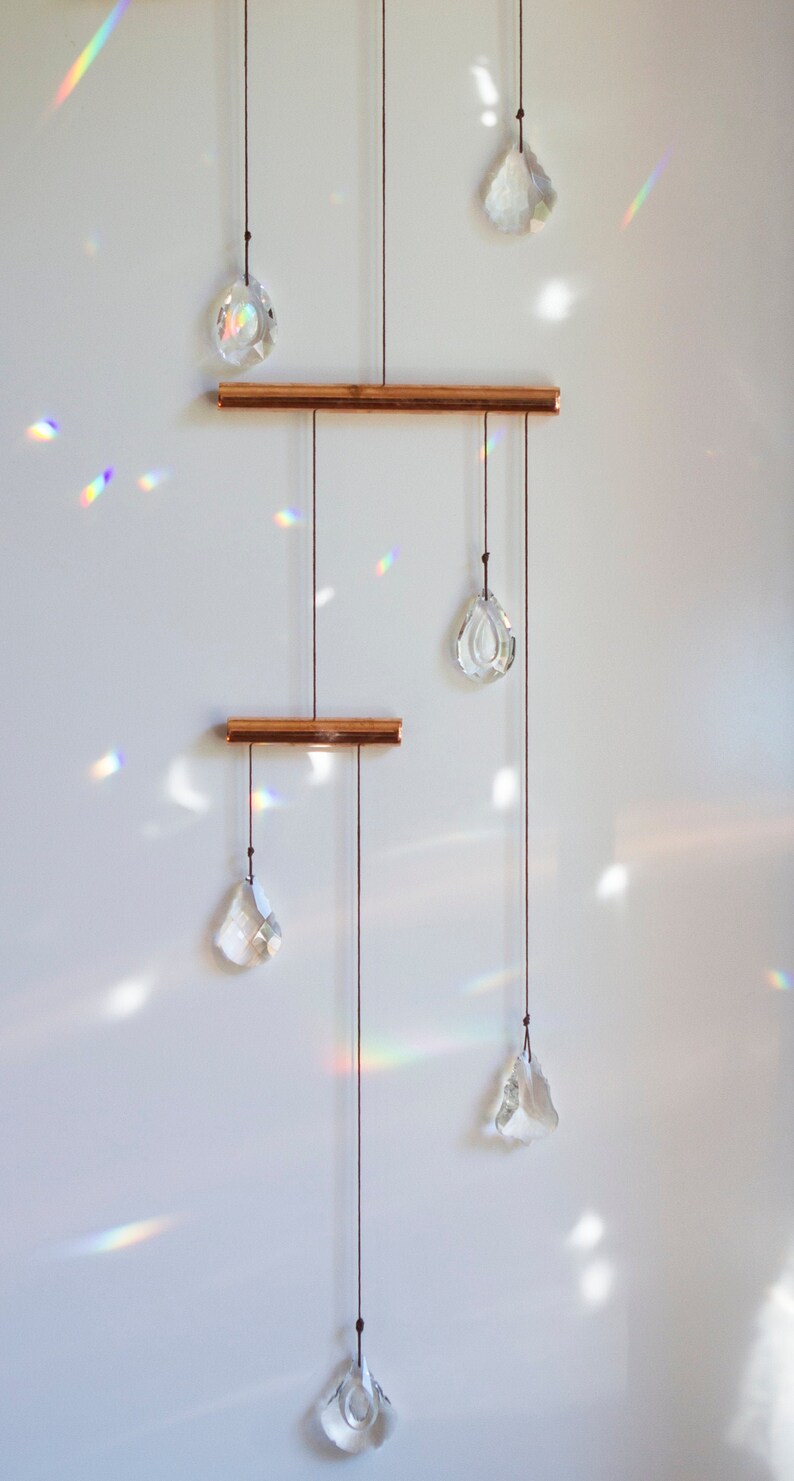 May include: A crystal prism mobile with six clear crystal prisms hanging from two copper bars. The prisms are suspended from strings and catch the light, creating rainbows on the white wall.