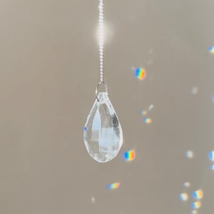 Peut inclure: Un pendentif en cristal clair et facetté en forme de goutte d'eau est suspendu à une chaîne argentée. Le cristal réfracte la lumière, créant de petits arcs-en-ciel. C'est un objet décoratif.
