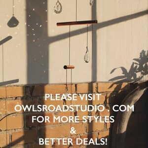 May include: A copper and crystal mobile with three teardrop-shaped crystals hanging from a copper bar. The mobile is hanging from a ceiling against a brick wall. The text "PLEASE VISIT OWLSROADSTUDIO.COM FOR MORE STYLES & BETTER DEALS! THANK YOU SO MUCH." is printed below the mobile.