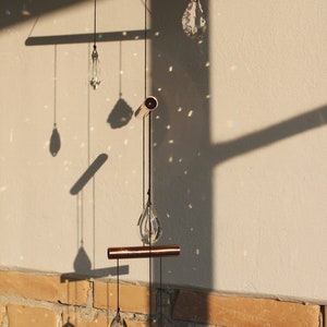 May include: A copper and crystal mobile with five teardrop-shaped crystals hanging from a copper bar. The mobile is suspended from the ceiling and casts rainbows on the wall behind it.