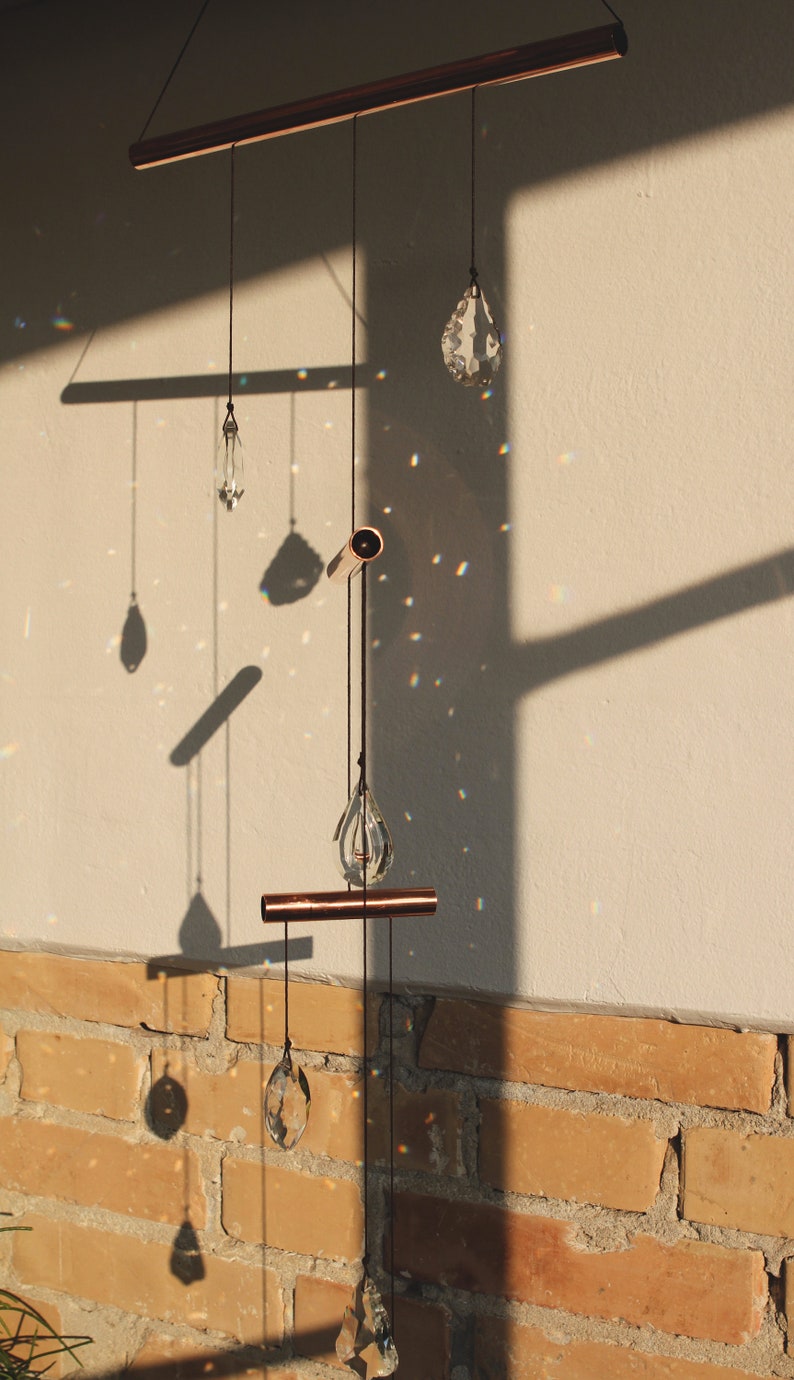 May include: A copper and crystal mobile with five teardrop-shaped crystals hanging from a copper bar. The mobile is suspended from the ceiling and casts rainbows on the wall behind it.