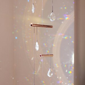 May include: A copper crystal suncatcher with five clear crystal drops hanging from it. The suncatcher is suspended from the ceiling and casts rainbows on the wall.