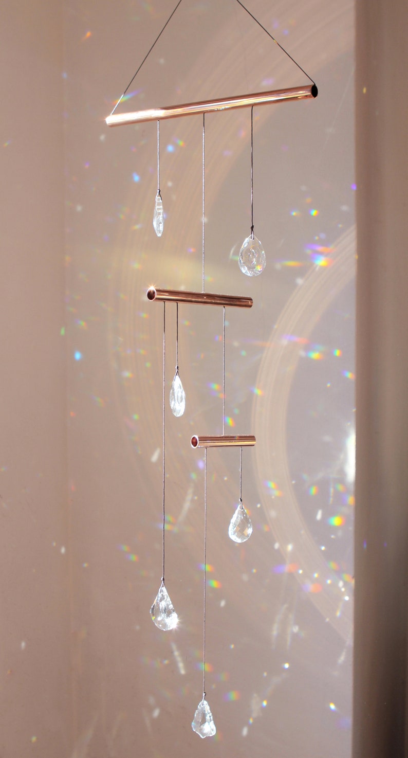 May include: A copper crystal suncatcher with five clear crystal drops hanging from it. The suncatcher is suspended from the ceiling and casts rainbows on the wall.