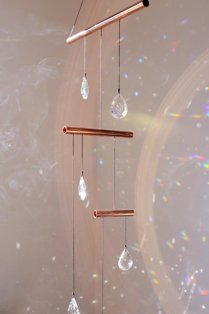 May include: A copper pipe mobile with five crystal drops hanging from it. The mobile is suspended from the ceiling and casts rainbows on the wall behind it.