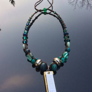 May include: A handcrafted necklace featuring a Hohner harmonica pendant. The necklace has a brown cord with turquoise and blue beads. The harmonica is silver and gold-toned. The necklace is designed to be worn.