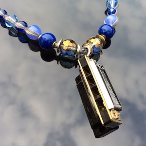 May include: A blue beaded necklace with a harmonica pendant. The necklace features various shades of blue beads, including round and faceted styles. The harmonica is black with gold-colored accents and a silver-colored plate.