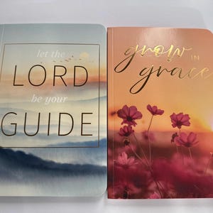 Könnte beinhalten: Zwei Hardcover-Journale. Eines zeigt den Text „Let the LORD be your GUIDE“ über einem Landschaftsbild. Das andere Journal hat den Text „grow in grace“ mit einem Blumen- und Sonnenuntergangsdesign. Beide Journale haben goldene Schrift.
