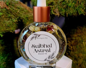 May include: A clear, round glass bottle filled with oil and dried botanicals, topped with a copper-colored collar and a white dropper. The label reads "The Pagans Way" and "Suibhal Astral Power Oil." It sits on a white hexagonal base.