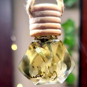 May include: A small, diamond-shaped glass bottle filled with a golden liquid and suspended by a thick, natural-colored jute rope. The bottle features a light-colored, ribbed wooden cap and contains visible botanical elements submerged in the liquid.