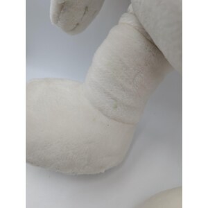 May include: Close-up of a white plush toy's leg and arm. The plush toy is made of a soft, fuzzy material. The leg is bent at the knee, and the arm is slightly raised. The background is plain white.