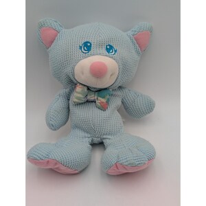 May include: A light blue teddy bear plush toy with pink accents on the ears, nose, and feet. The bear has embroidered blue eyes and a small bow tie with a striped pattern. The plush has a waffle-like texture.