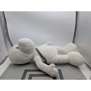 May include: A large, plush, white cartoon character with a round head, black eyes, and a gray strap across its body. The character is lying down on a gray cutting mat.