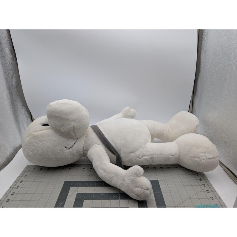May include: A large, plush, white cartoon character with a round head, black eyes, and a gray strap across its body. The character is lying down on a gray cutting mat.