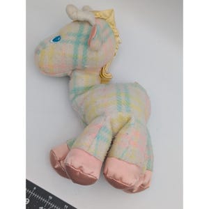May include: A plush toy horse in pastel colors. The horse has a plaid pattern in shades of pink, blue, and yellow. The hooves are a satin pink, and the mane and tail are a golden yellow. The horse has blue eyes and white ears.